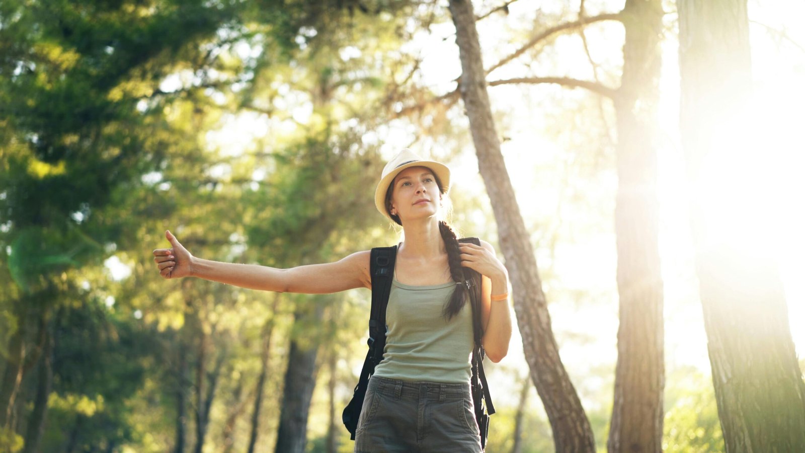 A woman with a hat and backpack hitchhiking through a sun-dappled forest.